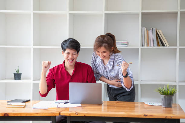 istock Cheerful business people using a laptop in an office. Happy young entrepreneurs smiling while working together in a modern workspace. Two young business people are sitting together at a table. 1702217274