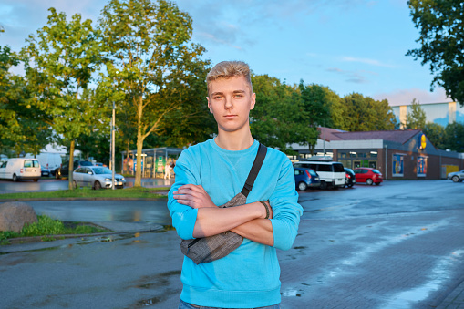 Portrait of handsome guy outdoor, on town street in sunset light. Young serious confident blond male looking at camera. Youth 18, 19 years old, lifestyle concept