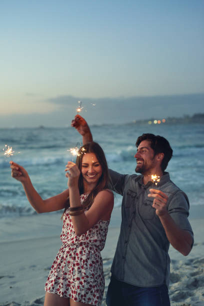 coppie felici che tengono scintille che festeggiano capodanno sulla spiaggia al tramonto con fuochi d'artificio scintillanti - 25 30 years foto e immagini stock