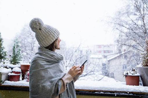 A woman in a warm hat and blanket holds smartphone in her hands. Winter landscape with snowfall in the background.