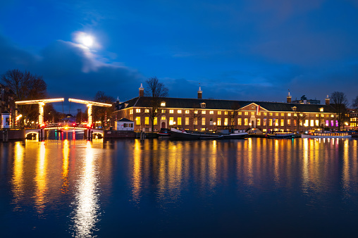 H'ART museum in Amsterdam with typical Amsterdam illuminated bridges at the river Amstel during a winter evening in the downtown canal district. The moon is shining in the dark evenin sky.