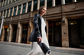 Portrait of blonde female fashion model in leather blazer jacket and white dress looking into the camera, walking in city. Young stylish woman outside