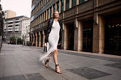 Full length fashionable blonde woman walking city street in chic attire with building in background. Young female model wears white fringed dress, black leather jacket and sandals, looks at camera