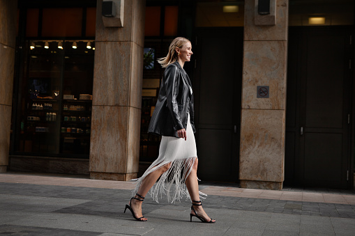 Full length happy blonde woman walking city street in stylish clothes. Young female model smiling, wears white fringed skirt, black leather jacket and sandals.