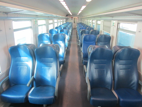 Empty train wagon. The blue leather seats are arranged in groups of two. Gray handrails for convenience. Large windows with roller shutters. Gray floor covering. Lamps for additional lighting.The sun's rays illuminate the interior of the train