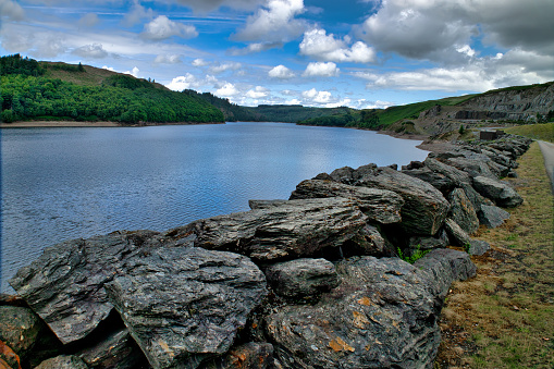 Llyn Brianne reservoir, Carmarthenshire, Wales, UK - While hiking, a view across the rocks lining the shoreline and the blue water of the reservoir toward forest surrounding the lake