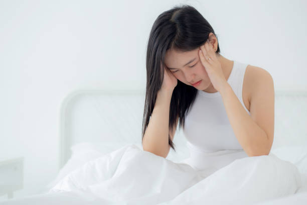 Young asian woman sitting on bed pain headache in the bedroom at home, unhappy female exhaustion and sick headache, anxiety and unwell, dizziness and disorder, medical and health concept. stock photo