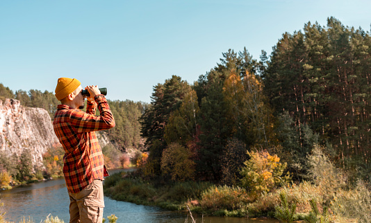 young man in red plaid shirt and yellow beanie hat looking through binoculars on river bank near cliff in fall Birdwatching and tourism copy space