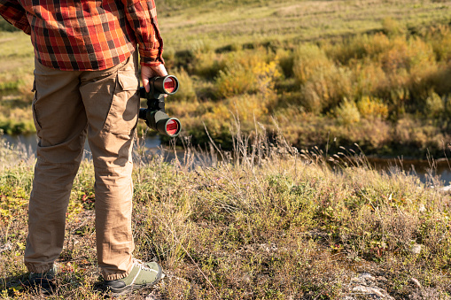 faceless man in red plaid shirt with binoculars standing on river bank near cliff in fall Birdwatching and tourism copy space