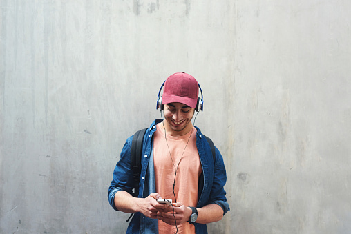 Portrait young man using smartphone listening to music wearing headphones with concrete wall background