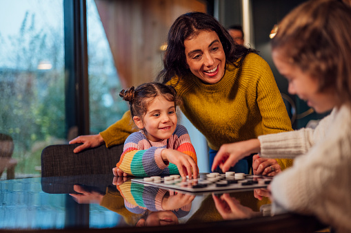 A medium shot of a mid-adult woman and her two daughters playing checkers on a winter's day in the North East of England. They are on a staycation holiday and staying in a luxurious holiday home.\n\n There is video to match this scenario.