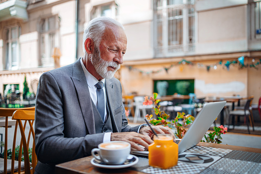 Cheerful senior Caucasian man sitting peacefully in a local cafe shop. He is working remote from his laptop and is enjoying his coffee and orange juice.