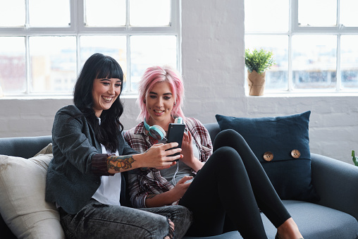 Two women friends using smartphones sitting on sofa at home watching entertainment on mobile phones