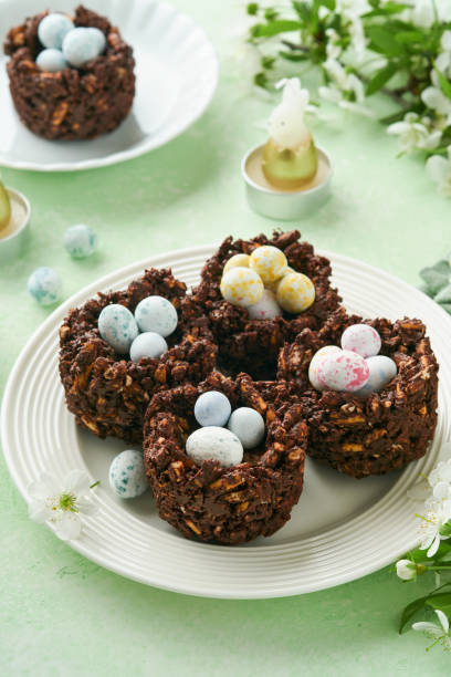 istock Easter chocolate nest cake with mini chocolate candy eggs with blossoming cherry or apple flowers on green background table. Creative recipe for Easter table with holiday decorations. Top view. 1702428363