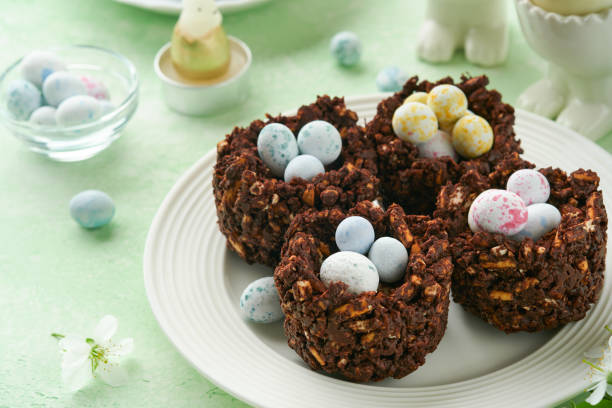 istock Easter chocolate nest cake with mini chocolate candy eggs with blossoming cherry or apple flowers on green background table. Creative recipe for Easter table with holiday decorations. Top view. 1702428909
