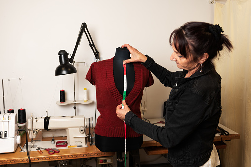 Woman is taking measures of a clothing on a mannequin with a tape measure in a sewing workshop. Sewing machine in the background