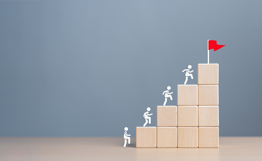 Business goal achievement and objective target for company success. Rises upstairs to Target. Human self-improvement and growth. Human icons climb on ladder stack block to the final goal red flag.
