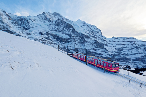On a sunny winter day, tourists travel on a cogwheel train from Jungfraujoch (Top of Europe) to Kleine Scheidegg on the snowy hillside with Jungfrau in background, in Berner Oberland, Switzerland