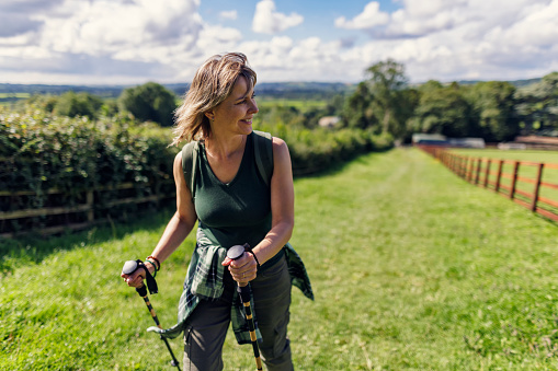 Single mature woman hiking in the Cotswolds, Gloucestershire, United Kingdom. She is walking with nordic walking poles on a green field.
Shot with Canon R5