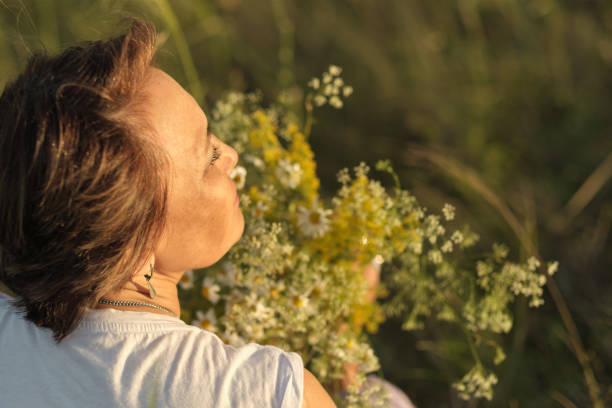thoughtful, modern retiree immerses herself in tranquility of summer meadow, wildflowers in her hands serving as poignant reminder of life's continuous journey and her own resilience through life una jubilada reflexiva y moderna se sumerge en la tranquilidad de la pradera de verano, con flores silvestres en sus manos que sirven como recordatorio conmovedor del viaje continuo de la vida y su propia resistencia a través de la vida.
