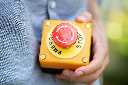 Man holding a large red industrial machinery emergency stop button in hand, object closeup detail, workplace occupational safety, security rules laws and policies, OHS work accidents simple concept