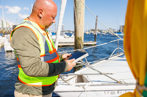 Mature bald appraiser in safety vest, with digital tablet inspects prow of yacht in pier in sunny day