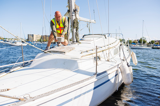 Ship prow evaluation: mature bald man in safety vest, with digital tab inspects yacht prow, assessing damage and facilitating insurance claim processing in pier