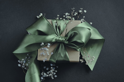 Paper gift box with olive green ribbon tied in a bow, small flowers, black background. Decoration for holidays, birthdays, weddings.