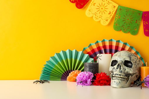 Set mood with side view shot of table decked out in Day of Dead theme. Tableau showcases macabre calavera skull, vivid fans, flowers, candles, spiders, garland on yellow wall, space for text or promo