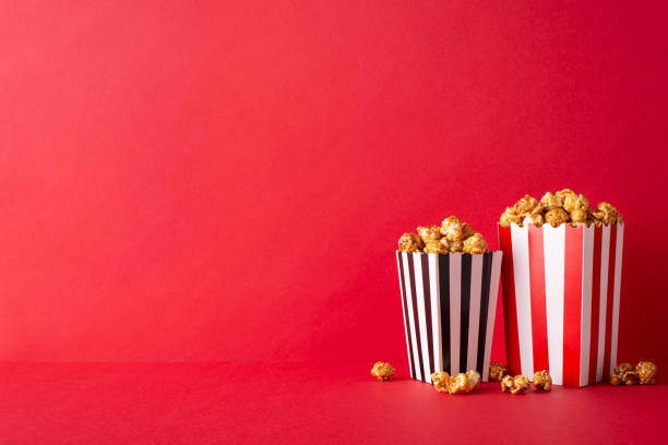 istock Cinematic delight at its best: A side view table stacked with cheese and caramel popcorn in striped boxes, against a red wall, ready for a movie premiere with friends 1702909342