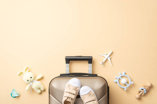Newborn's first flight: Top view of small plane model, suitcase, pacifier, knitted bunny toy, wooden rattle, teether, and tiny boots against pastel beige backdrop, great space for your promo message