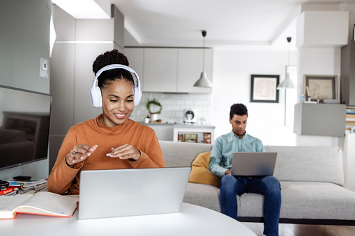 Young multiracial couple working at home in the same room. They are concentrating on the work. A woman with wireless headphones is sitting at the table and having a video call on the laptop. A man is sitting on the couch with a laptop on his lap.