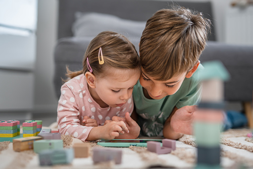 Two children brother and sister laying on the floor at home and playing games on mobile phone, early child development concept