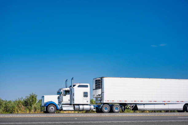 istock Clean powerful classic white big rig semi truck tractor transporting cargo in refrigerator semi trailer driving on the highway road with green line on the side 1703193434
