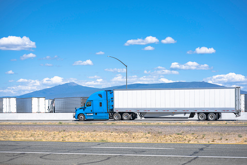 Industrial grade loaded long hauler carrier blue big rig semi truck tractor transporting commercial cargo in dry van semi trailer running on the highway road drives past a truck stop in California