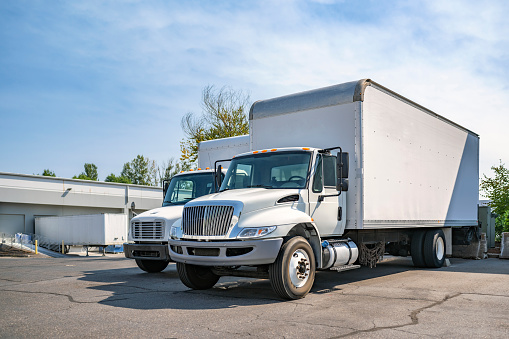 Middle duty Industrial standard white day cab rig semi trucks with box trailers for local deliveries and small business needs standing on the warehouse parking lot waiting for the next load