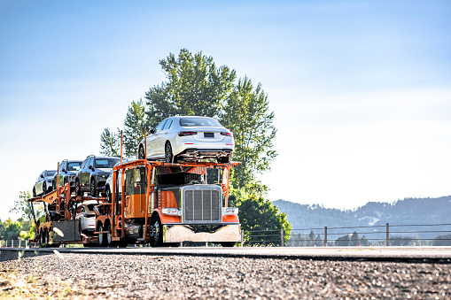 Bright orange industrial standard car hauler classic carrier big rig semi truck tractor transporting cars two level hydraulic modular semi trailer driving on the divided highway road in California