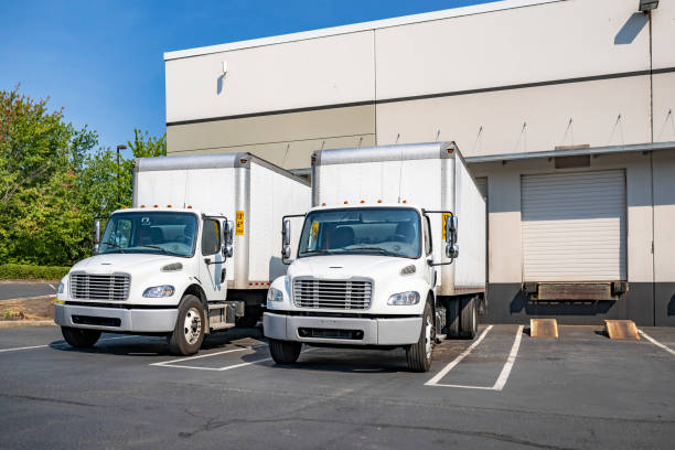istock Two compact middle day cab rig semi trucks with box trailers standing on the warehouse parking lot with dock and gates loading cargo for the next freight 1703228020