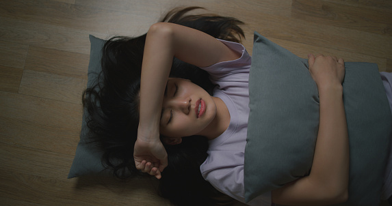 Portrait of Depressed young Asian woman hugging pillow while lying on the floor in the living room at home. Female lost in thoughts, and suffer from psychological. Mental health and people concept.
