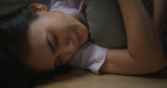 Portrait of Depressed young Asian woman hugging pillow while lying on the floor in the living room at home. Female lost in thoughts, and suffer from psychological. Mental health and people concept.