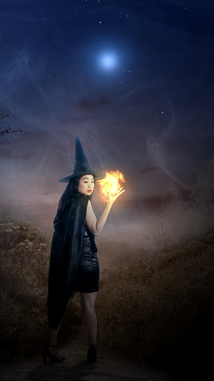 Scary witch showing magic spell in her hand in the misty hillside in the night. Scary witch for Halloween. Halloween concept