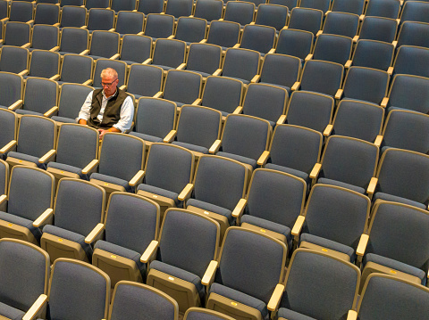 Overhead view a middle age man with gray hair sitting alone in an empty, auditorium seats, chairs.