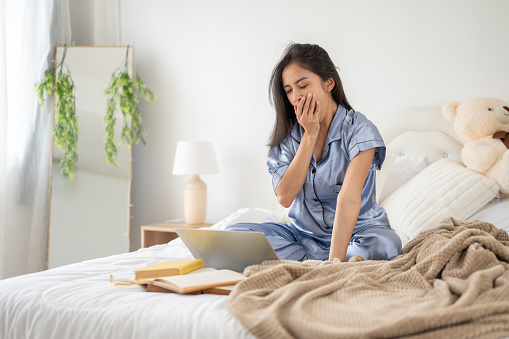 A sleepy and tired Asian woman in pajamas is covering her mouth and yawning while working on her laptop on her bed, feeling overworked.