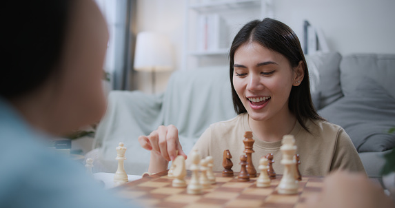 Selective focus, young woman moving pieces on chess board during competition with friend, they are talking and smile with fun during challenge in chess game together
