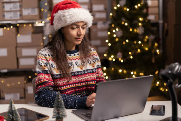 istock Salesman in a Santa Claus hat takes an order by laptop computer while sitting in warehouse with boxes for Christmas. Shopping and delivery. Small business 1703452041