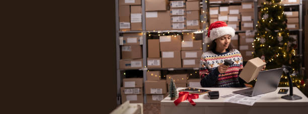 istock Scanning barcode on delivery parcel. Worker scan barcode of cardboard packages on Christmas holiday. Woman in Santa hat working in warehouse scanning labels on the boxes with barcode scanner. 1703452043