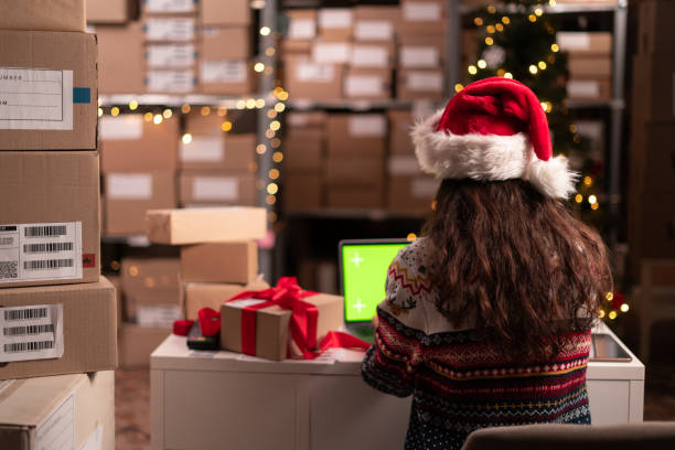 istock Female manager in Santa hat using green mockup screen laptop computer to check inventory. Warehouse retail center with boxes on christmas sale, e-Commerce Online Orders 1703452058