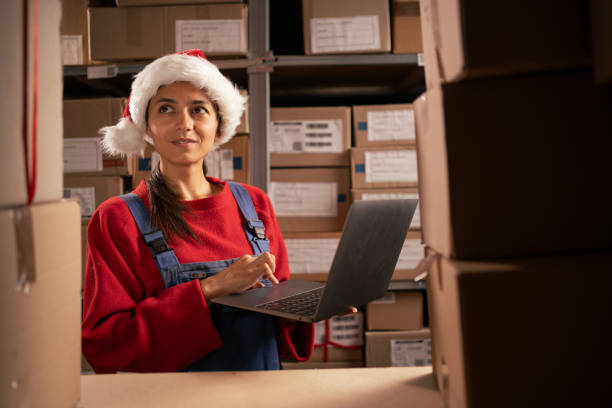 istock Worker checking inventory using Laptop Computer. Young female Santa Claus working in a warehouse with rows of shelves with parcels and orders ready for shipment. 1703470913