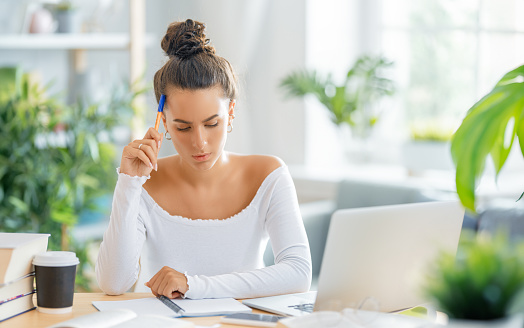 Focused young businesswoman studying online, watching webinar, podcast on laptop, making notes, sitting at work desk. E-learning concept.