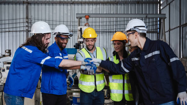 Team engineers and foreman stack hand and shake hands to show success at factory machines. Workers are controlling welding robots.